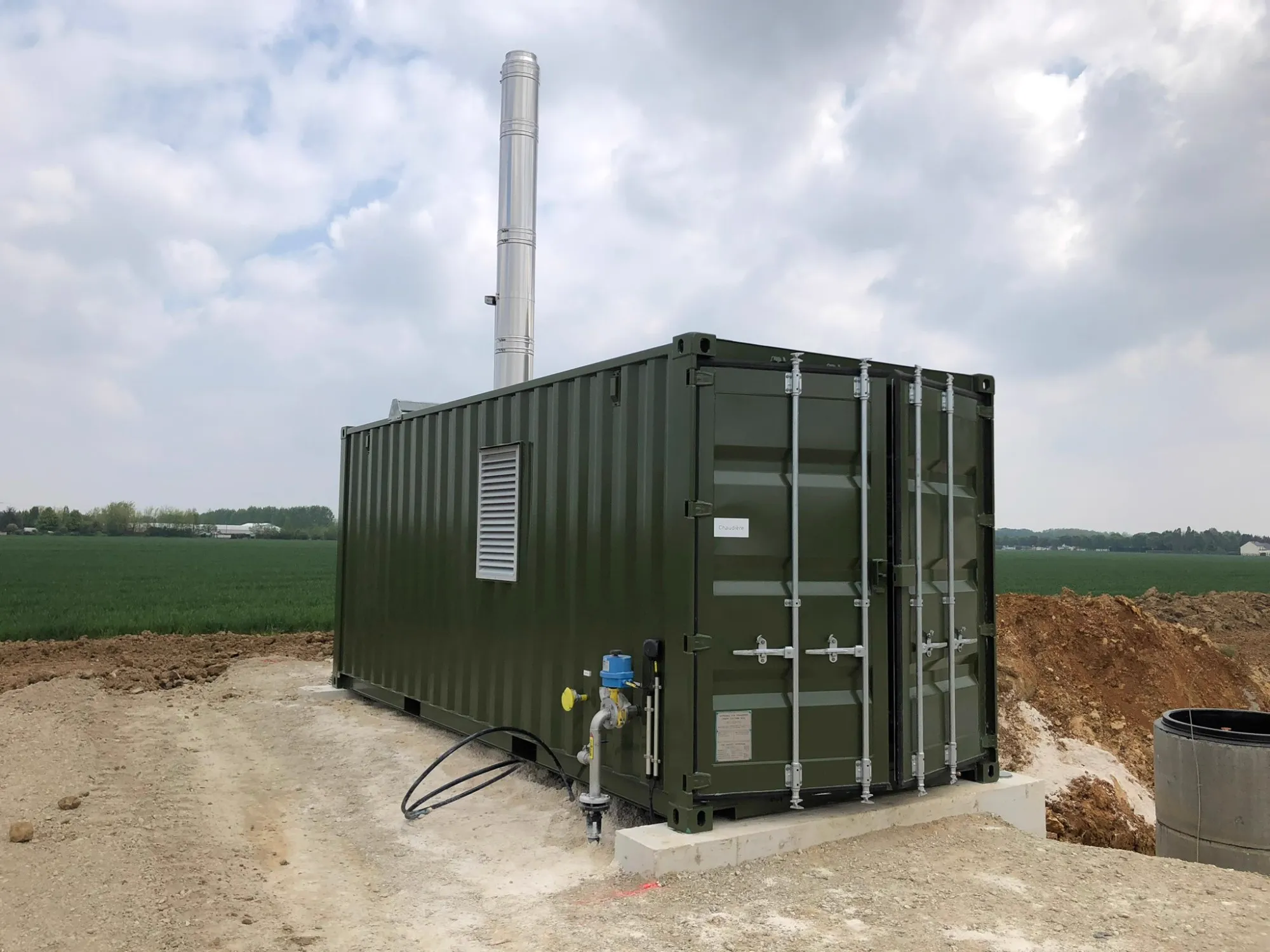 Grüner HeizMax-Heizcontainer mit Schornstein auf einem Fundament im Feld